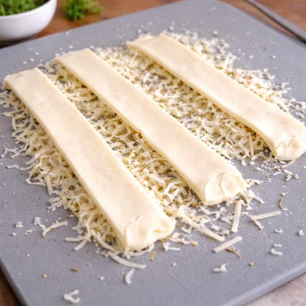 four long strips of raw puff pastry laid out on a grey baking tray. Underneath and around the strips there’s a generous layer of grated cheese, with visible herbs