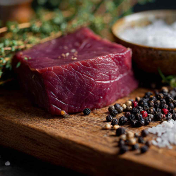 Raw venison steak on a wooden cutting board, surrounded by fresh thyme, rosemary, crushed juniper berries, black peppercorns and bay leaves, a small ceramic bowl with coarse sea salt,